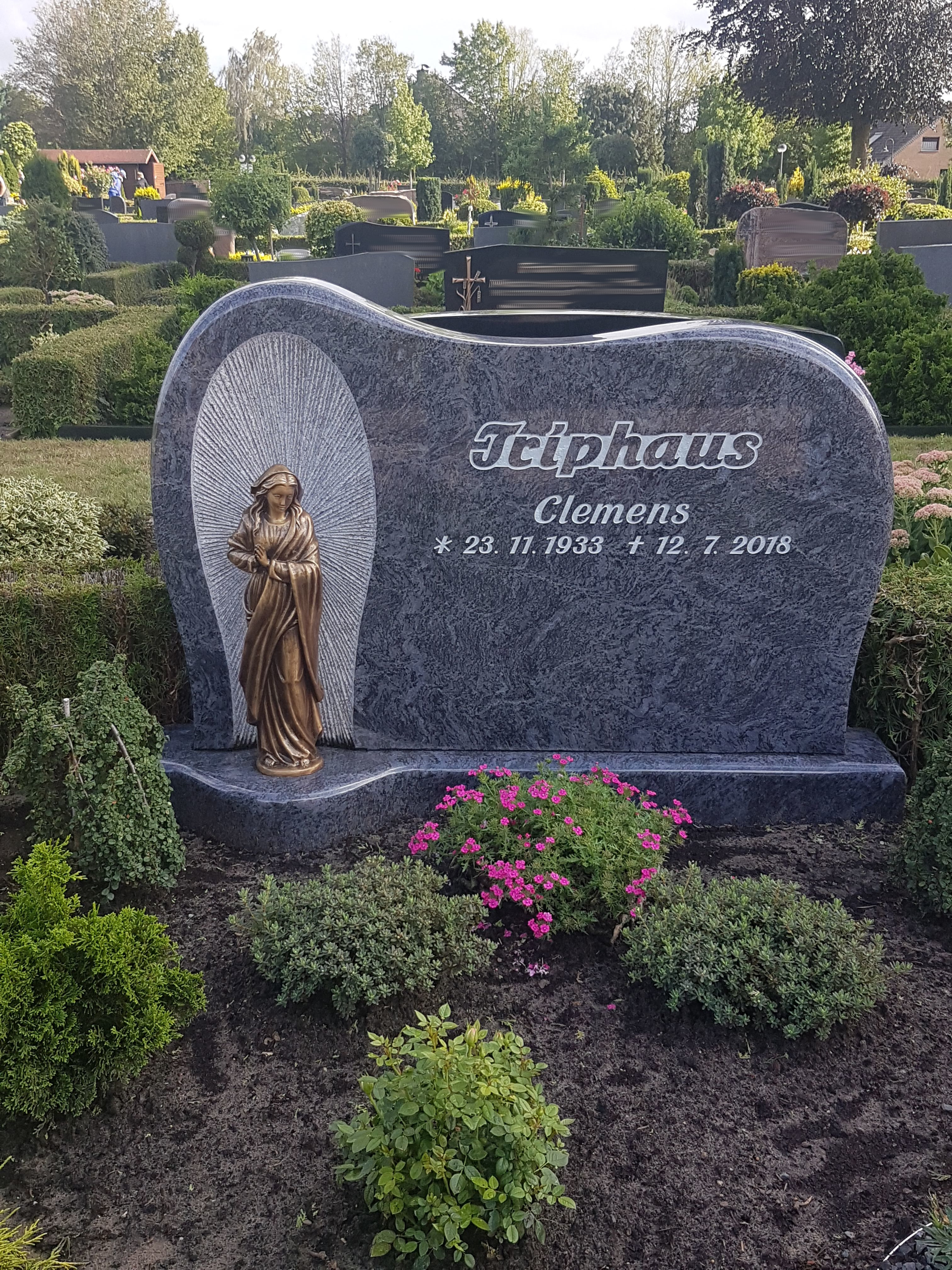 Ein grauer Grabstein mit goldener Heiligenfigur und edler Inschrift im grünen Garten