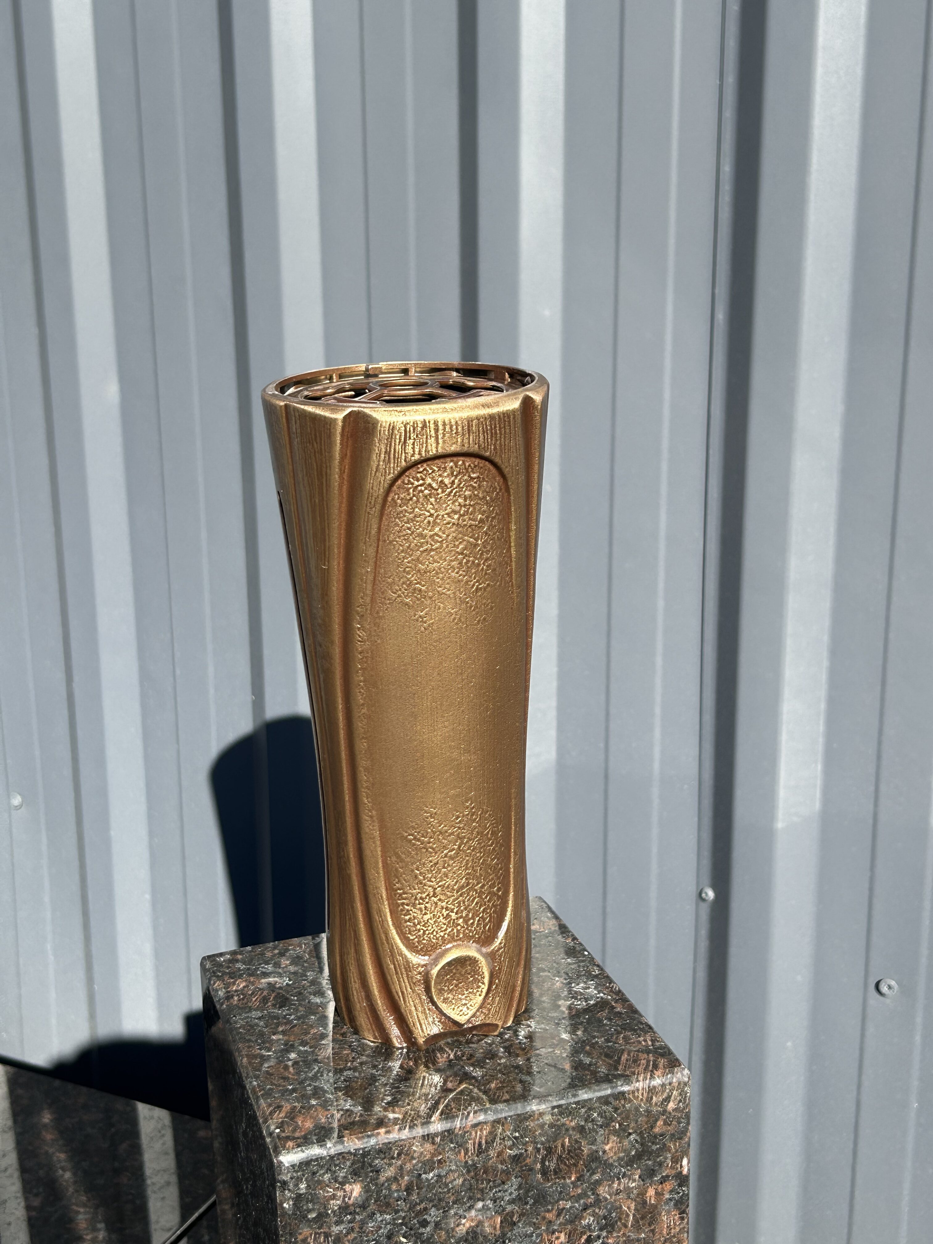 Eine goldene, säulenförmige Skulptur mit strukturierten Flächen steht auf einem dunklen Marmorsockel vor einer grauen Metallwand und glänzt in der Sonne