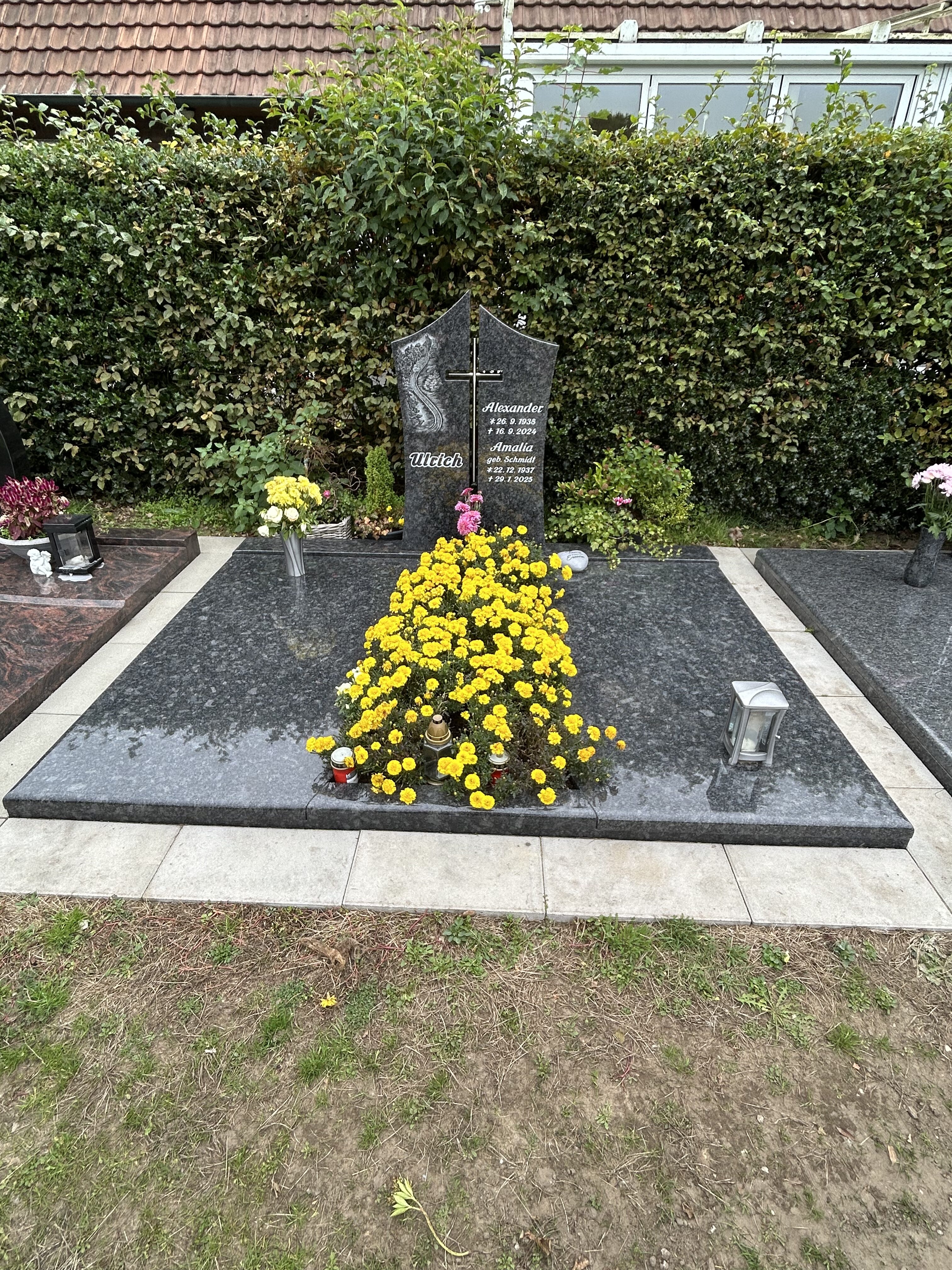 Ein schwarzgrauer Grabstein mit Kreuz und Blumen, bedeckt von gelben Chrysanthemen