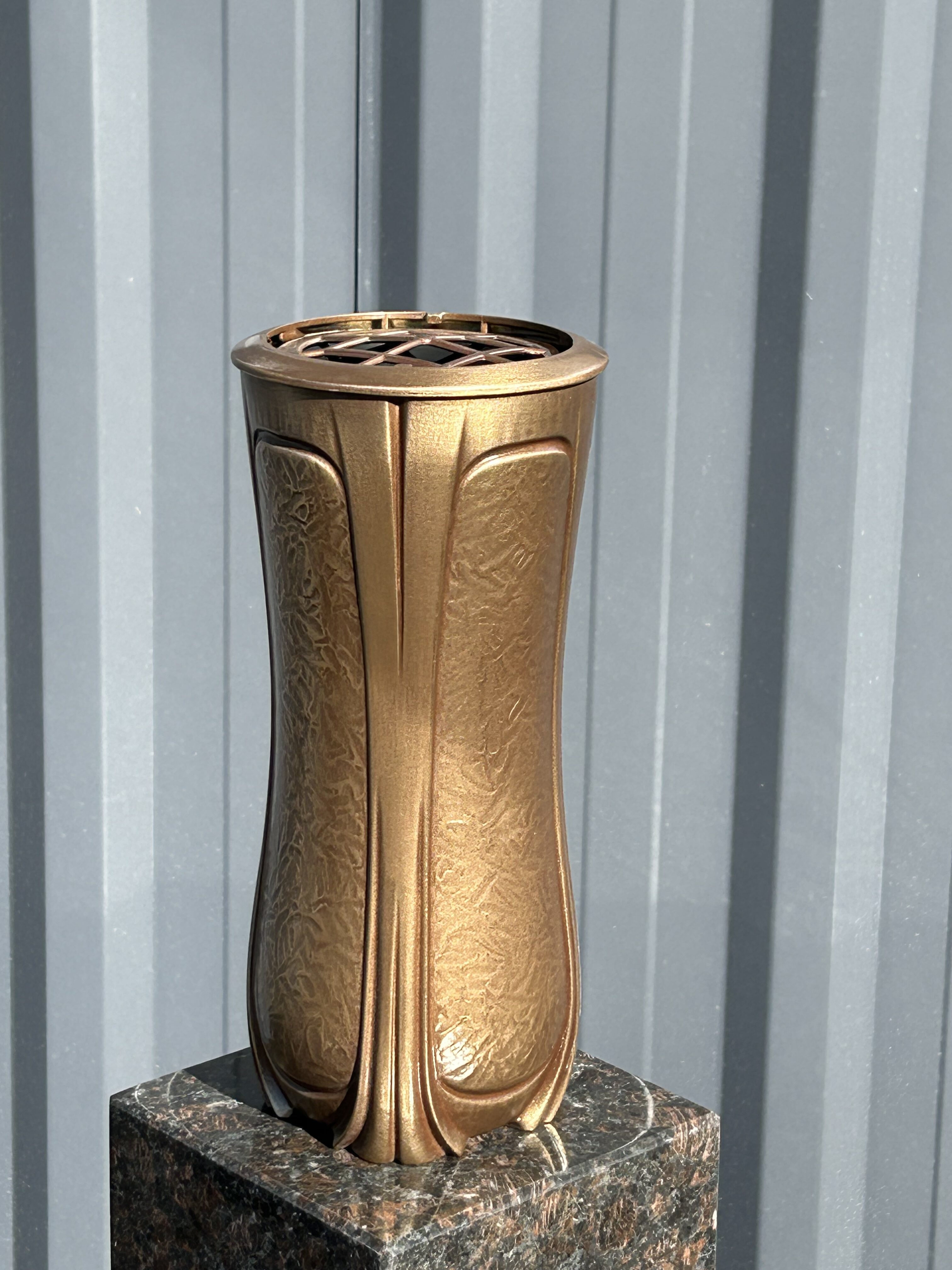 Eine bronzefarbene Vase mit floralen Mustern steht auf einem Granitsockel im Sonnenlicht