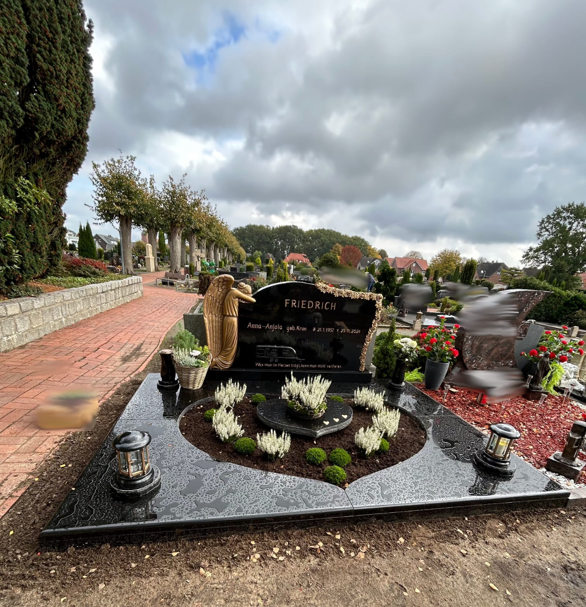 Ein schwarzer Grabstein mit goldener Engelsfigur steht auf einem gepflegten Friedhof