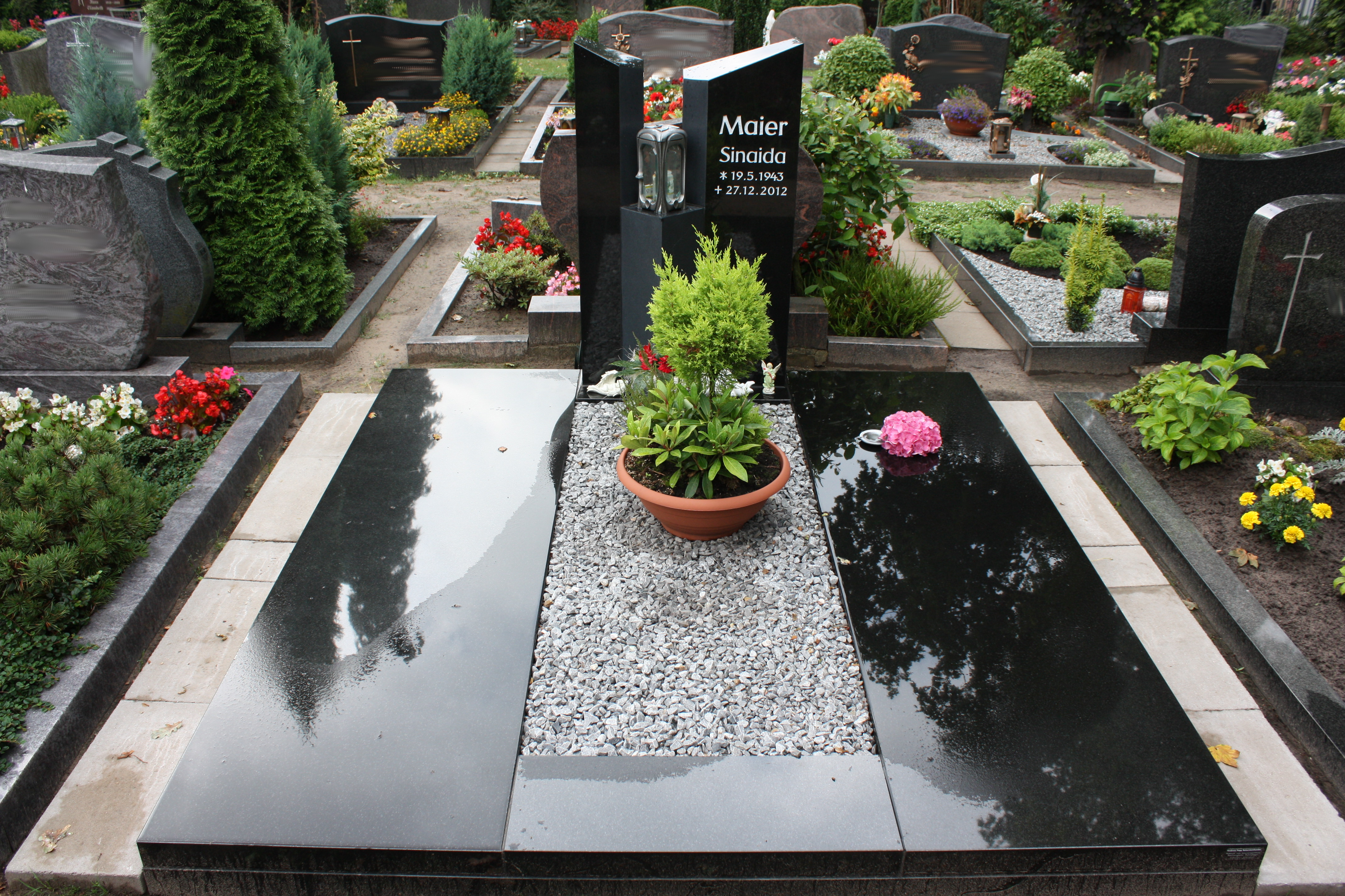 Ein gepflegtes Grab mit schwarzem Stein, hellem Kies und Blumen in einem Topf