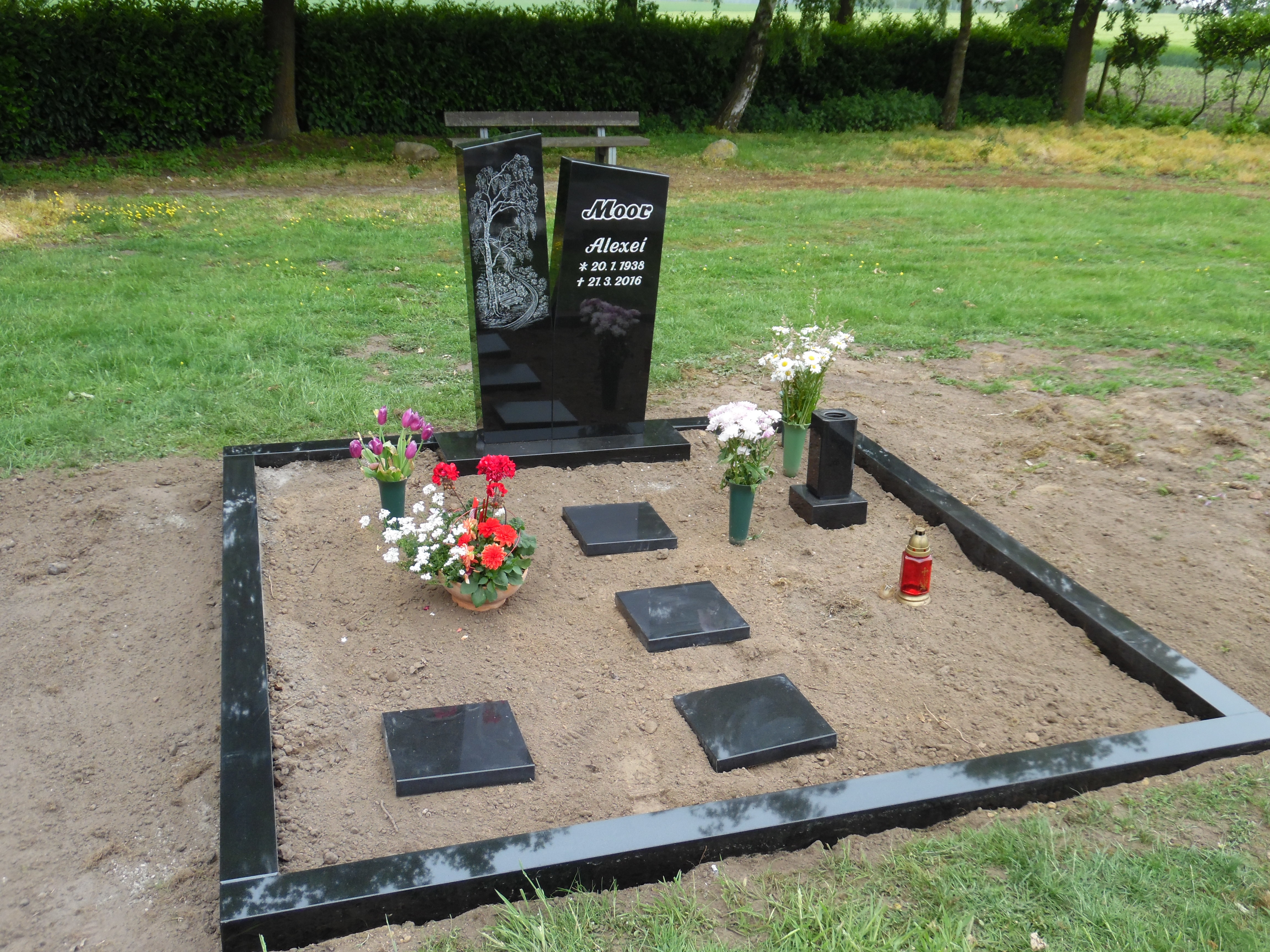 Schwarzer Grabstein mit eingraviertem Engel und Blumen, umgeben von Erde und Rasen