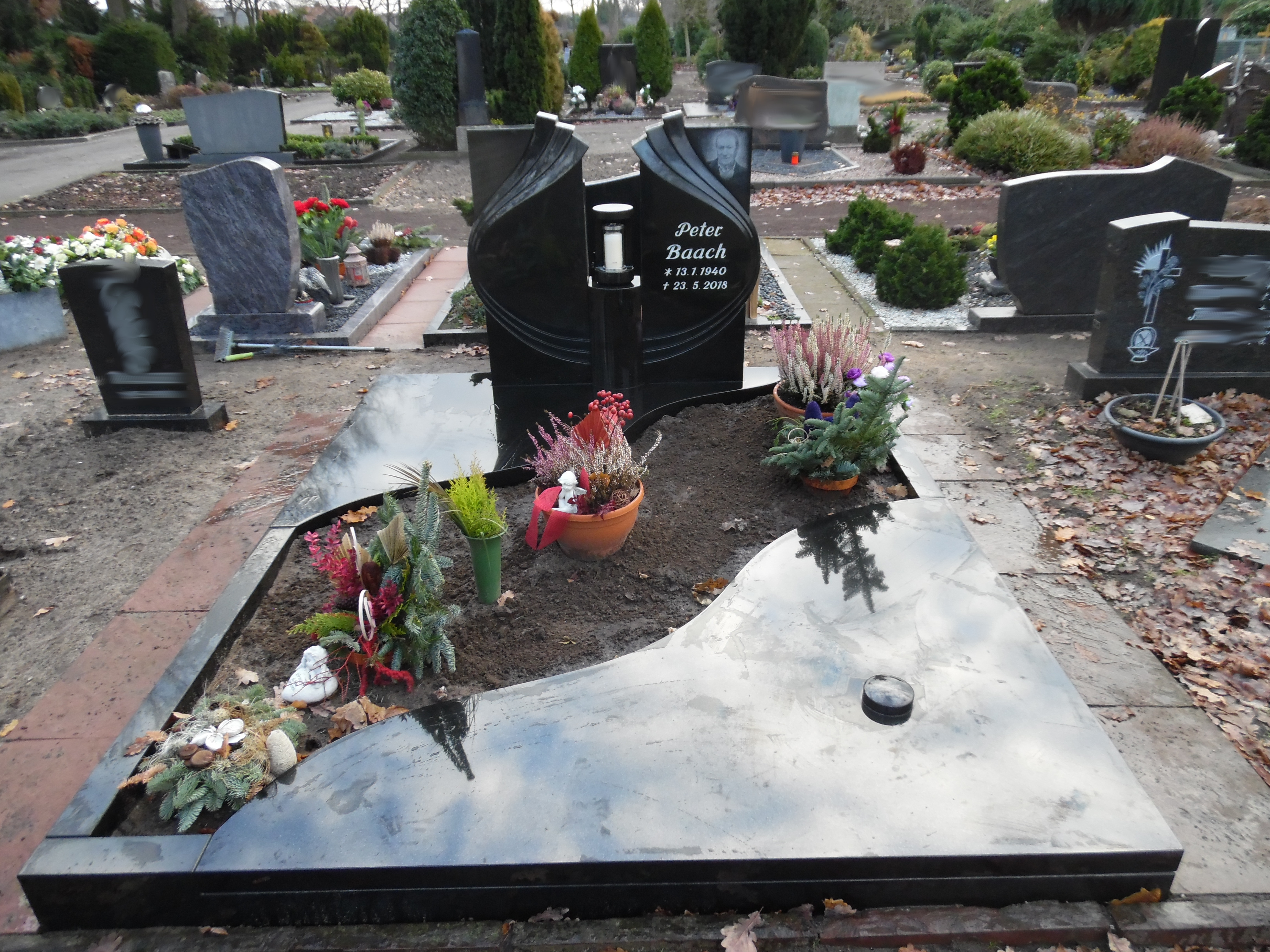 Schwarzer Grabstein mit dem Namen Peter Basch, Blumen und Pflanzen schmücken das Grab
